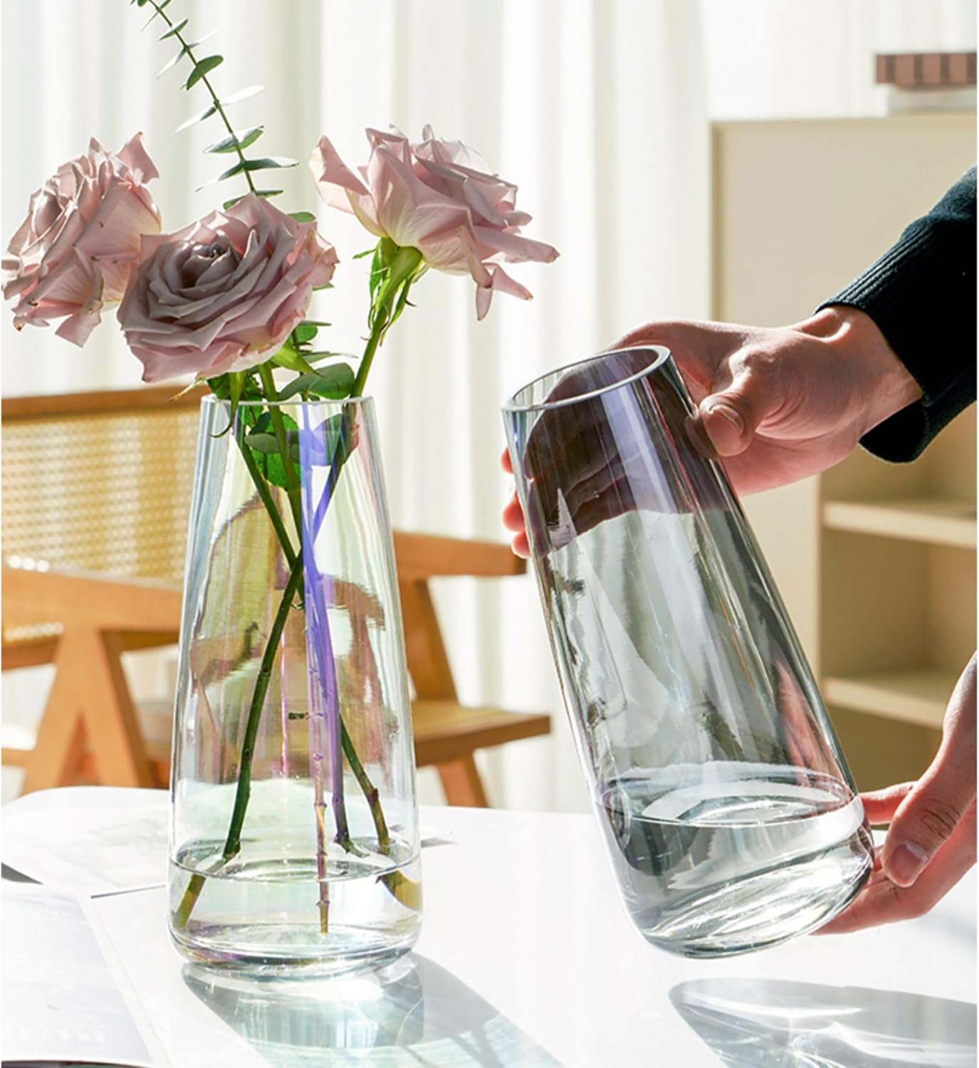 Clear glass vase with pink roses on a table, person holding another clear glass vase.