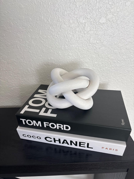 Decorative knot sculpture on top of two books titled 'Tom Ford' and 'Coco Chanel' on a white wall background.