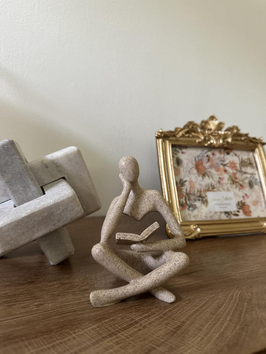 Decorative sculpture of a sitting figure on a wooden surface with a framed picture in the background.