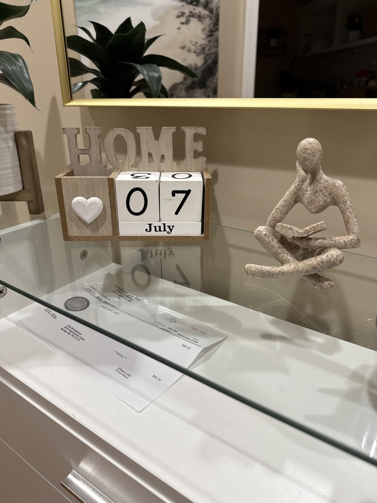 Decorative setup with a calendar showing July 7th, a 'HOME' sign, and a small statue on a reflective surface.