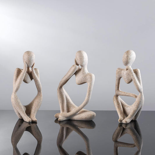 Three stone sculptures of abstract human figures on a reflective surface with a light gray background