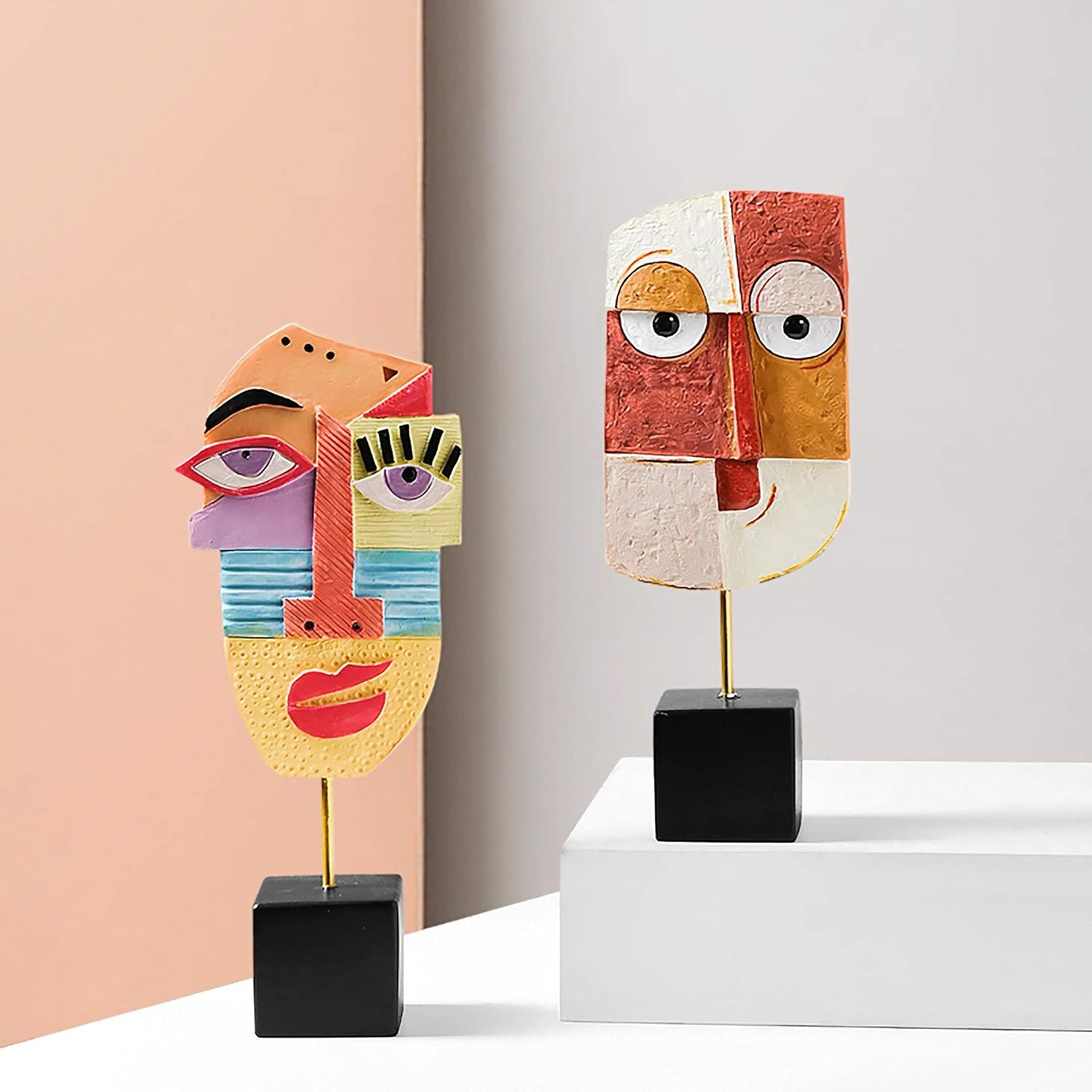 Two abstract face sculptures on stands against a light pink and white background