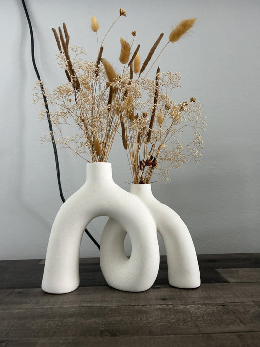 Two white, abstract vases with dried plants on a wooden surface.