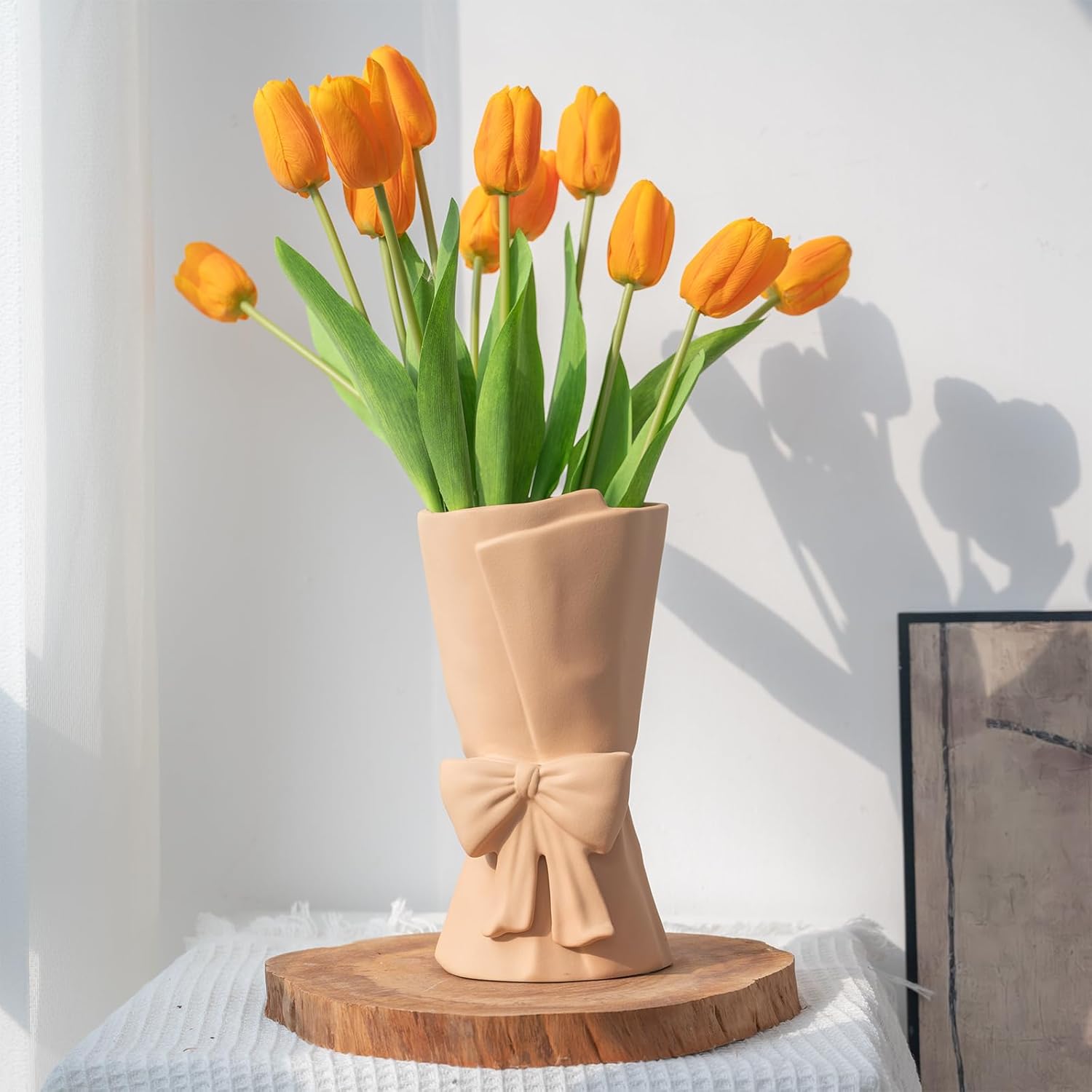 Beige vase with a bow design holding orange tulips on a wooden stand against a white wall.