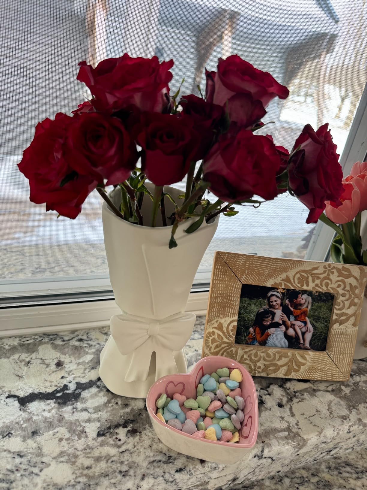 Bouquet of red roses in a white vase on a marble surface with a heart-shaped container of candies and a framed photo.
