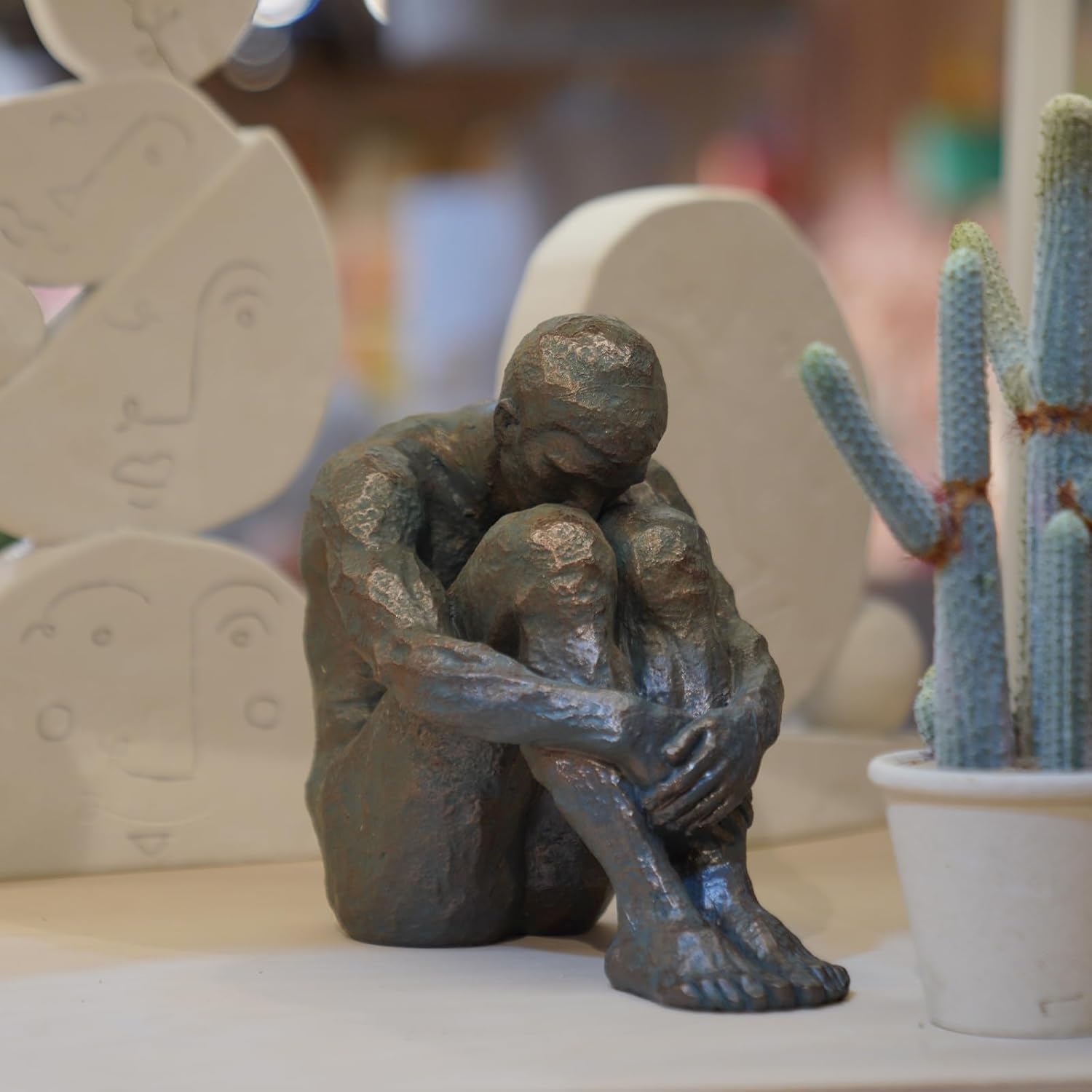 Bronze sculpture of a person sitting with hands on knees, blurred background with cactus and abstract shapes.