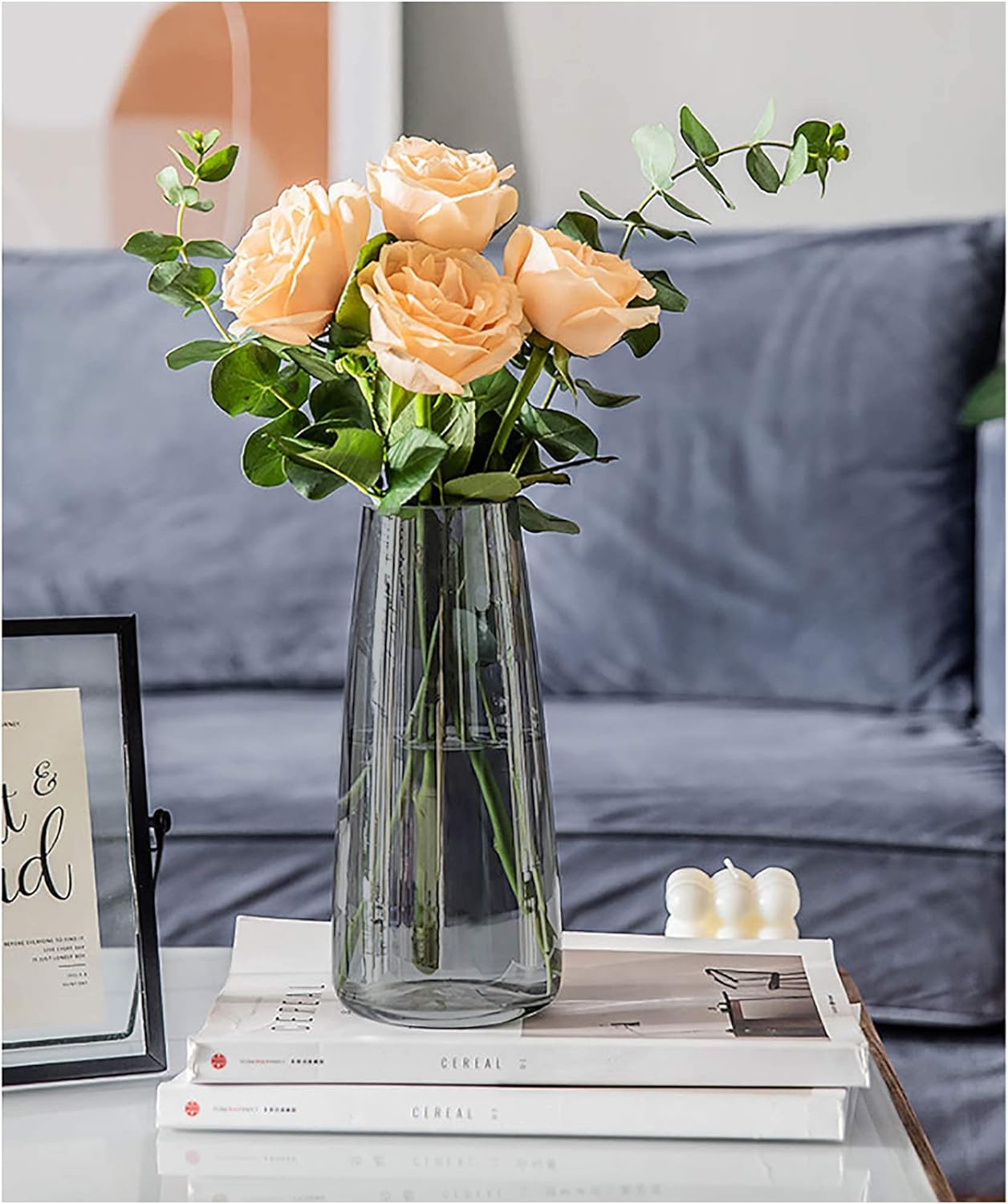 Clear vase with peach-colored roses on a table with books and candles, against a blurred sofa background