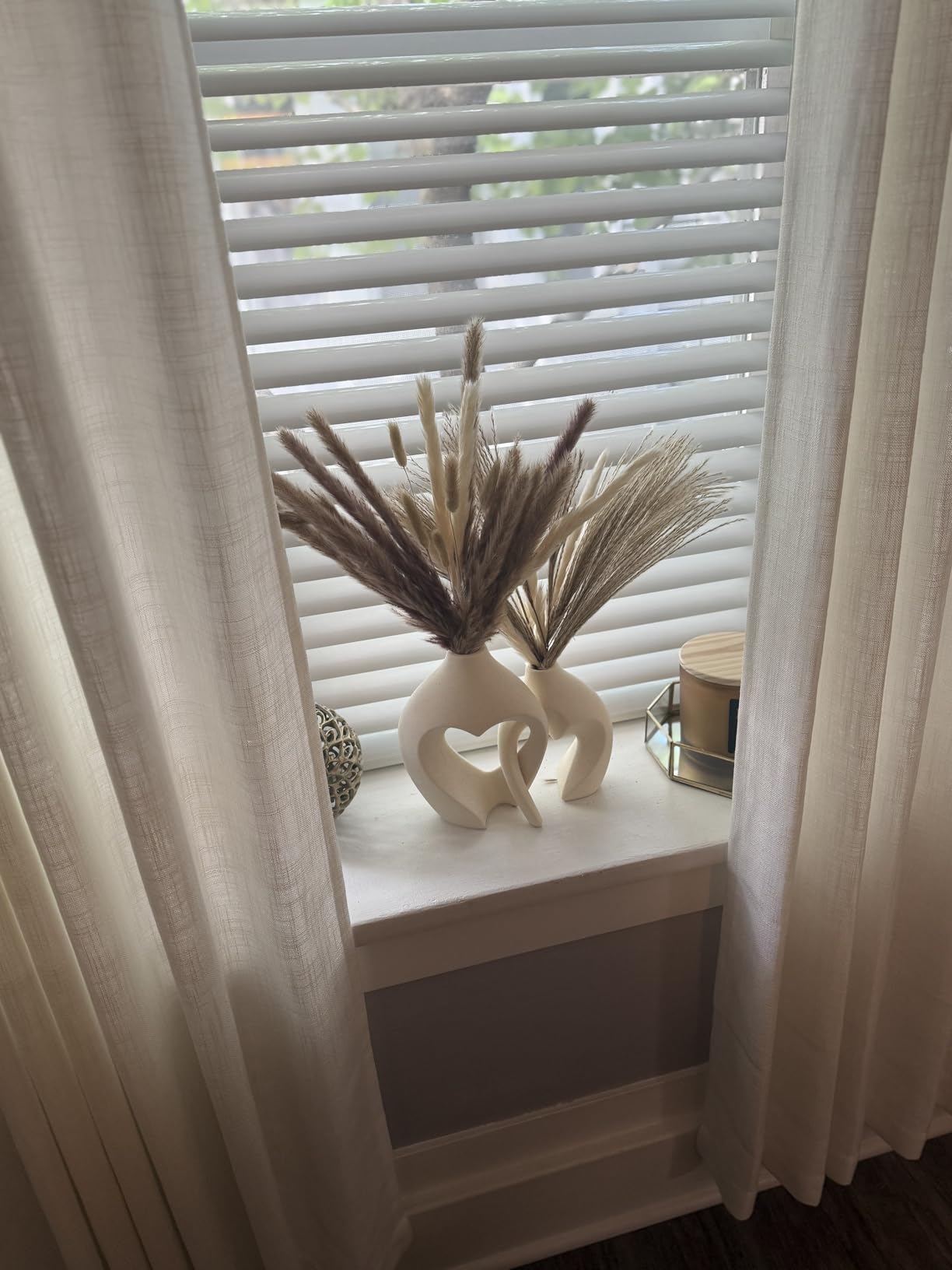 Decorative vase with dried plants on a window sill with light filtering through curtains.