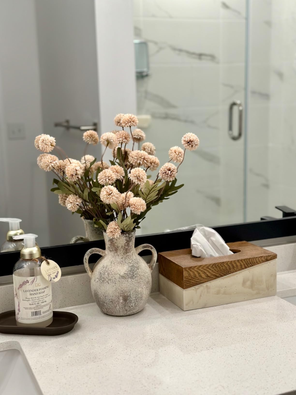 Decorative vase with flowers on a bathroom counter next to a tissue box and bottle.