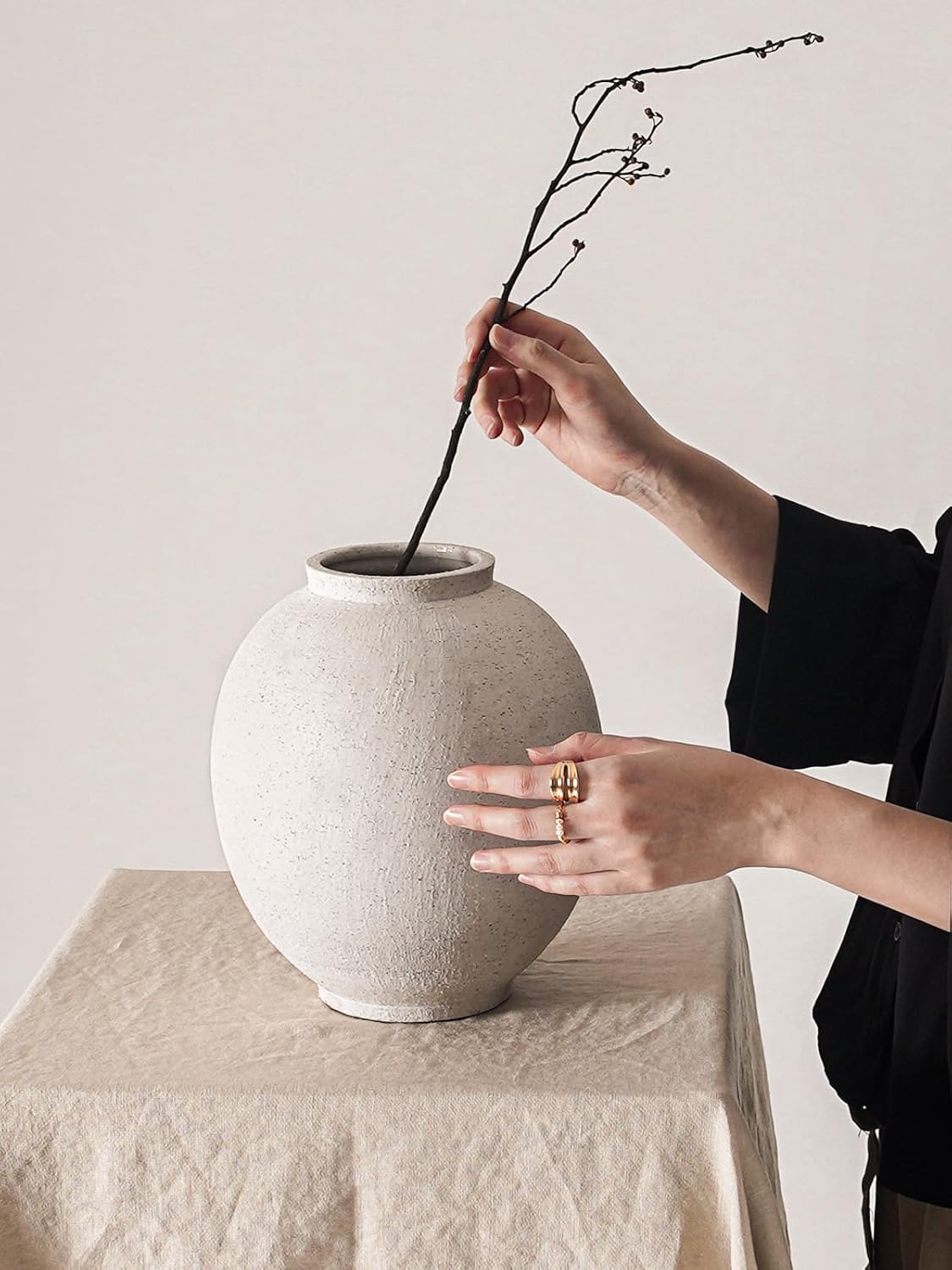 Person arranging a branch into a textured ceramic vase on a neutral surface.