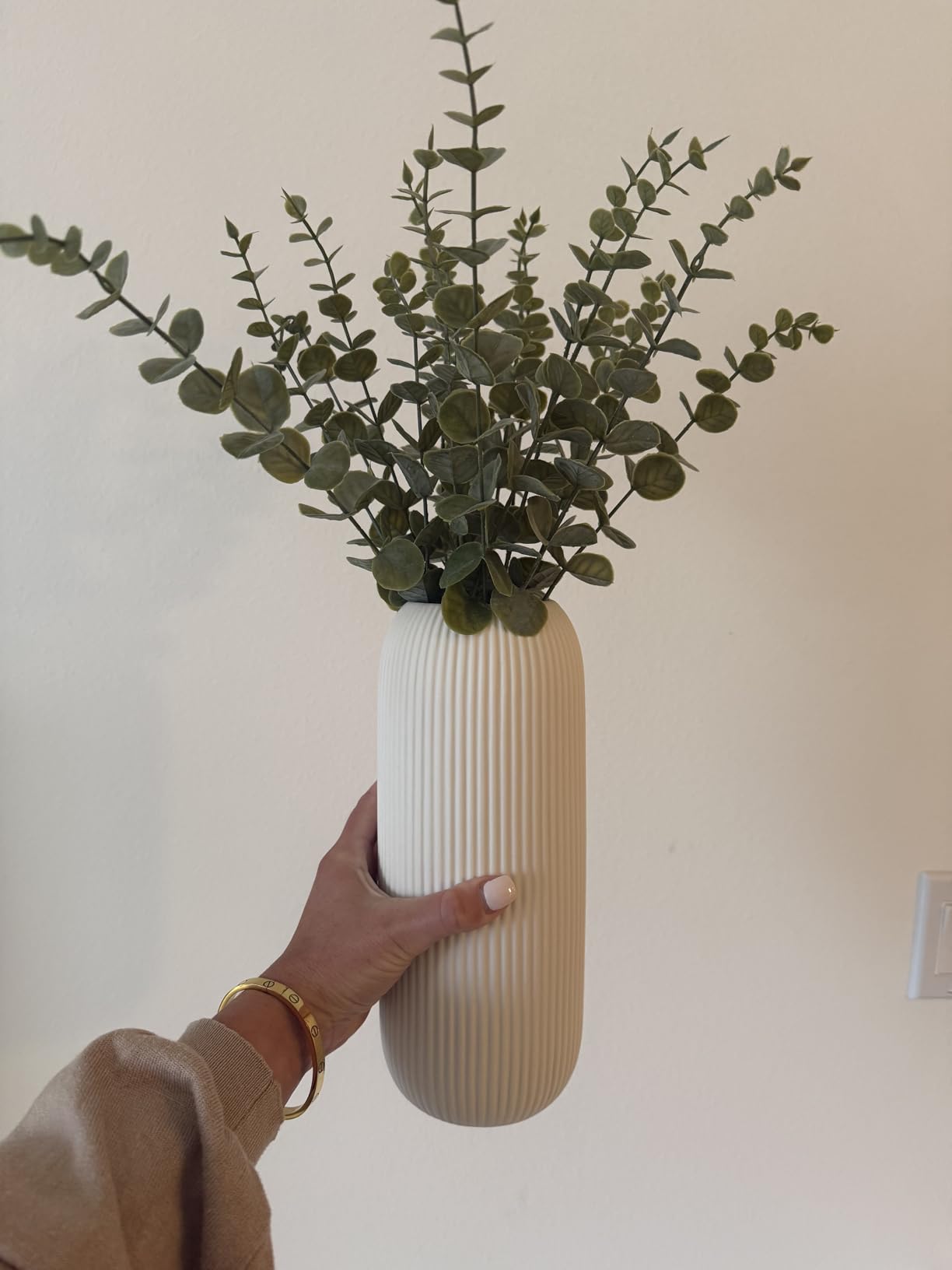 Person holding a textured white vase with greenery against a plain wall