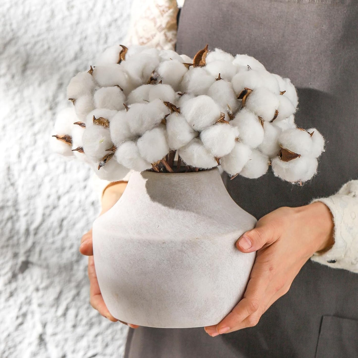Person holding a white vase with cotton-like flowers against a textured gray background
