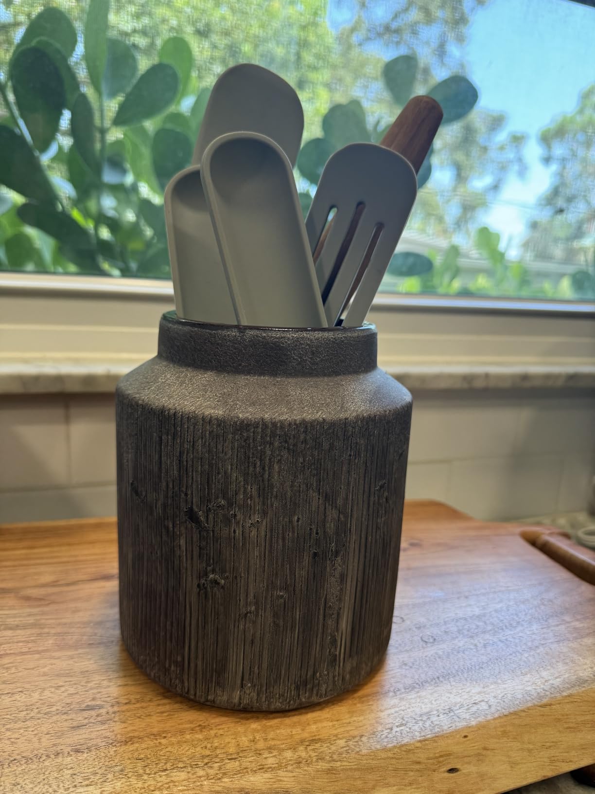 Set of kitchen utensils in a wooden holder on a wooden surface with a window in the background.