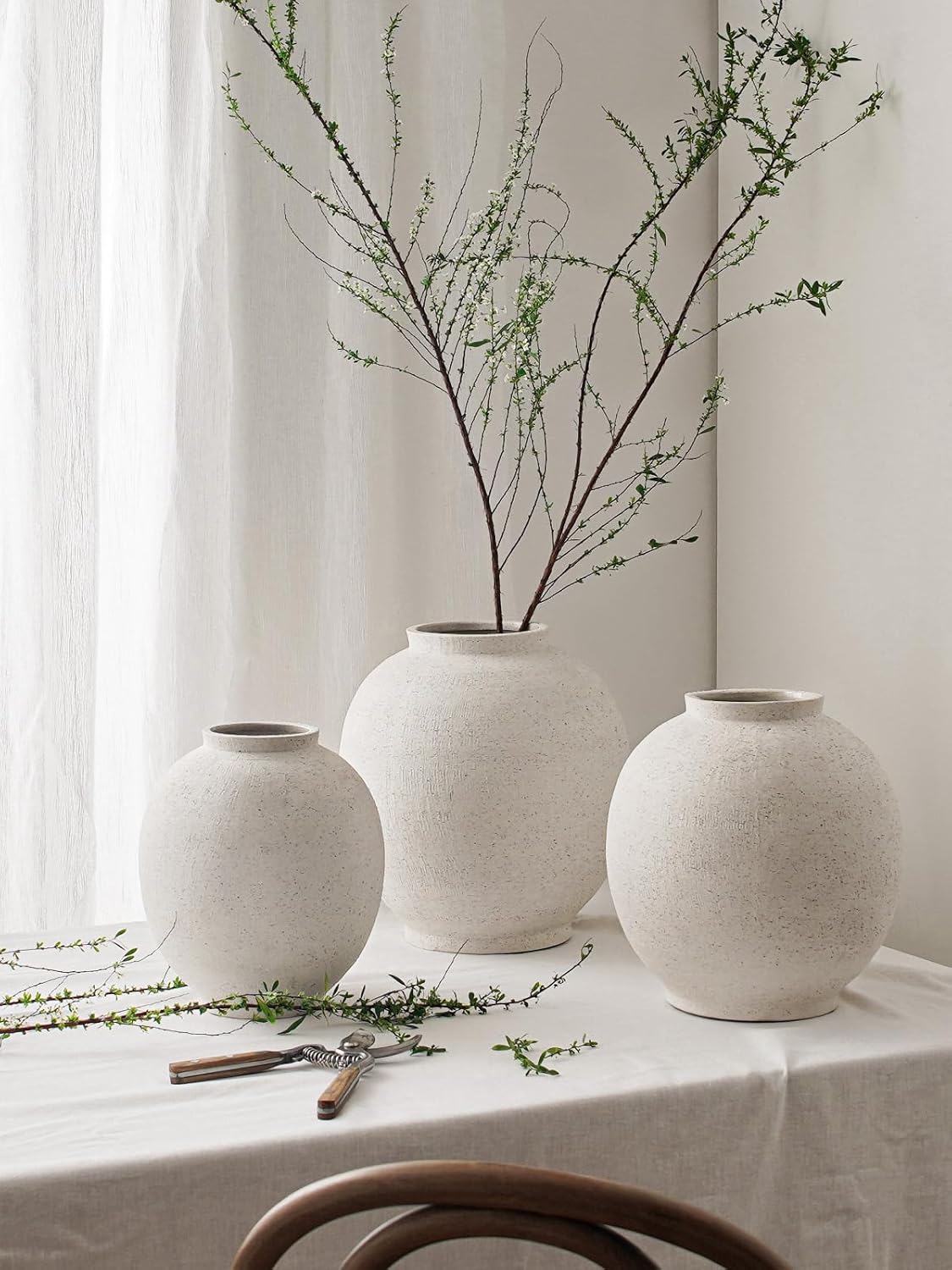 Three textured stone vases on a table with greenery against a white curtain.