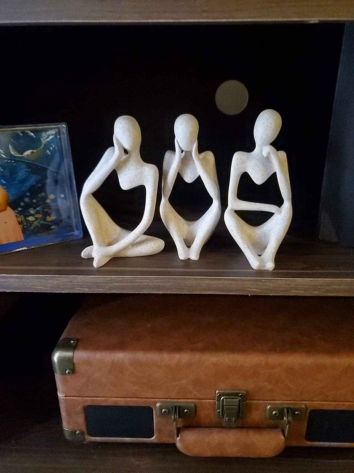 Three white ceramic figures on a shelf above an open brown suitcase.
