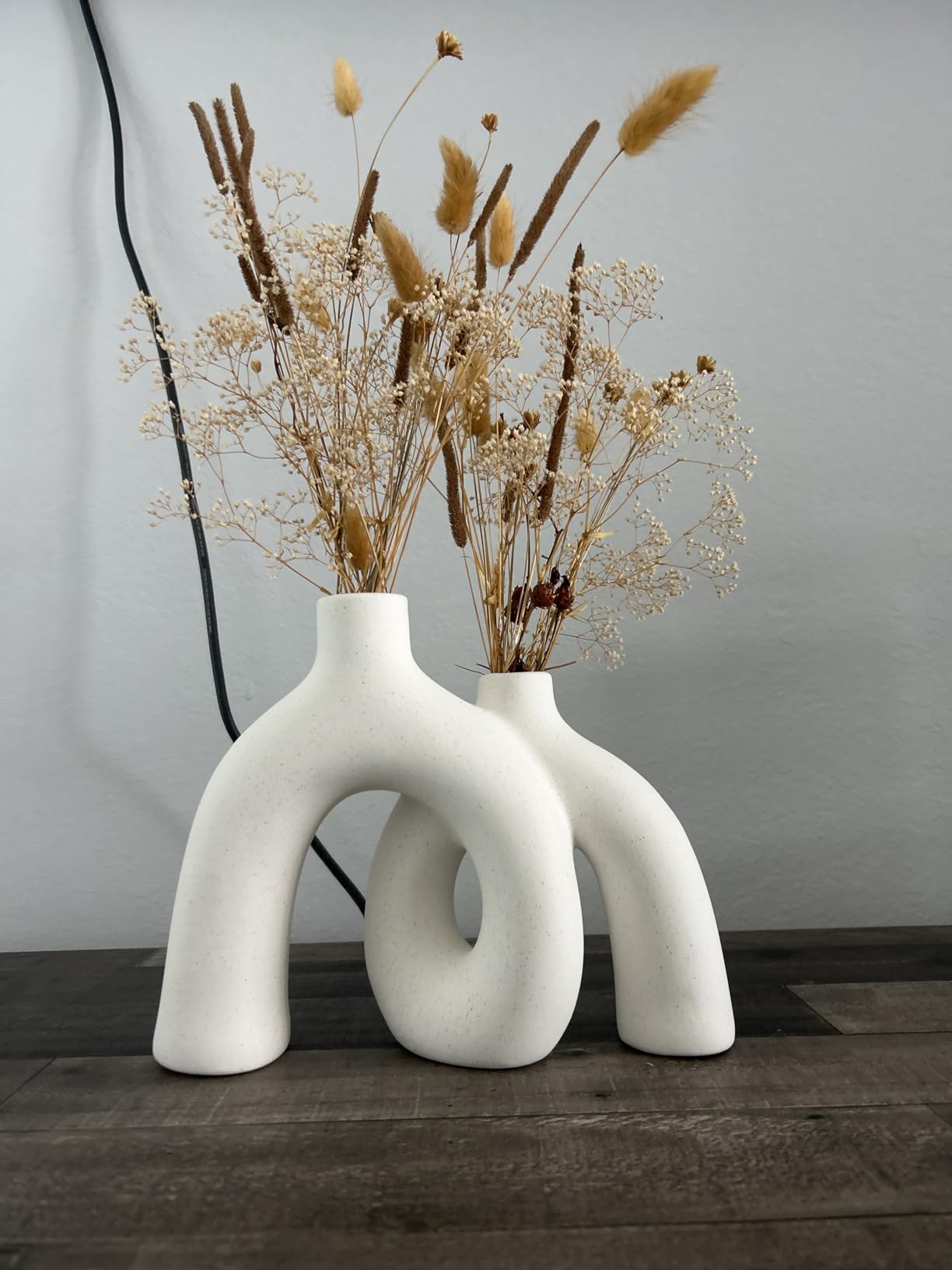Two white, abstract vases with dried plants on a wooden surface.