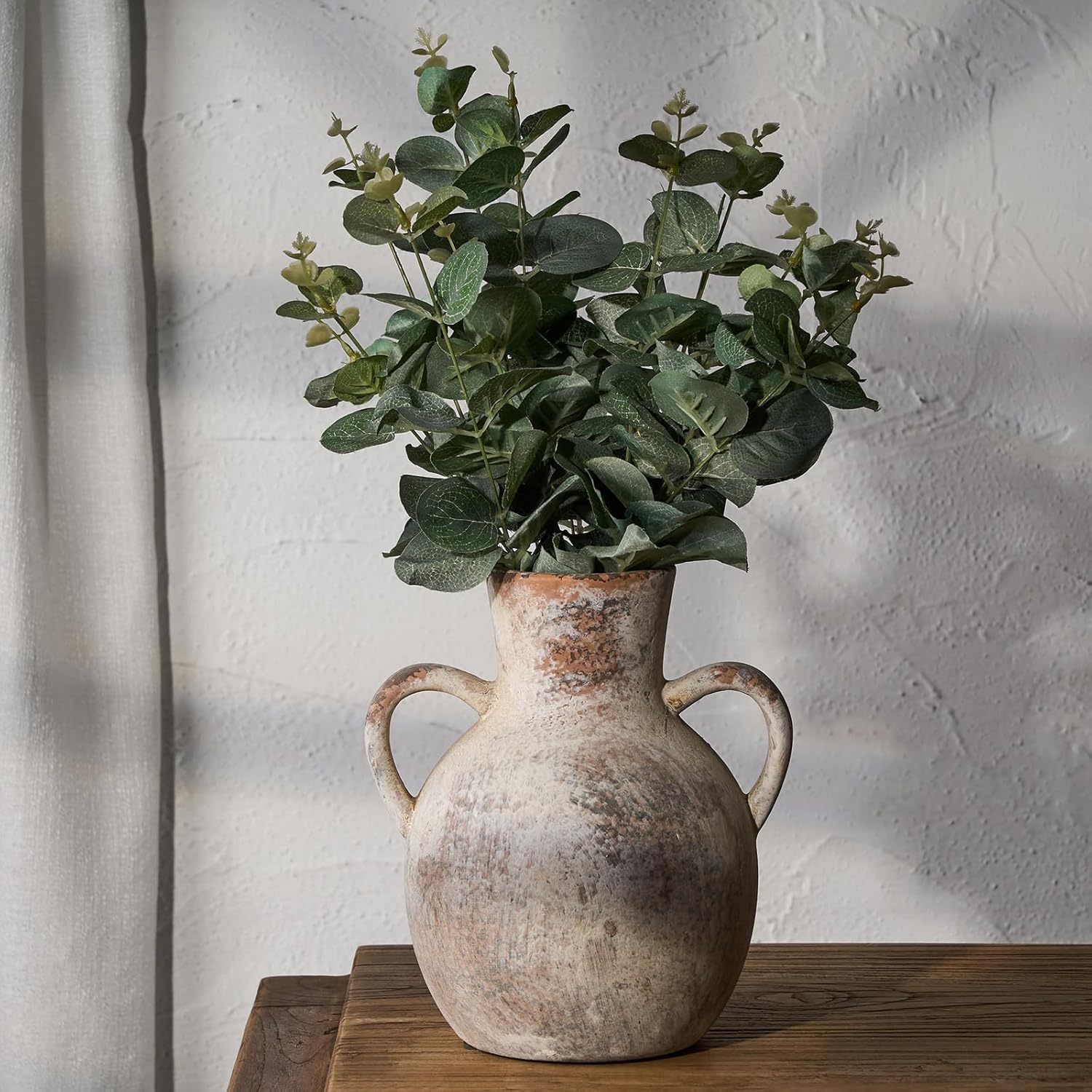 Vintage-style vase with eucalyptus on a wooden surface against a textured white wall.