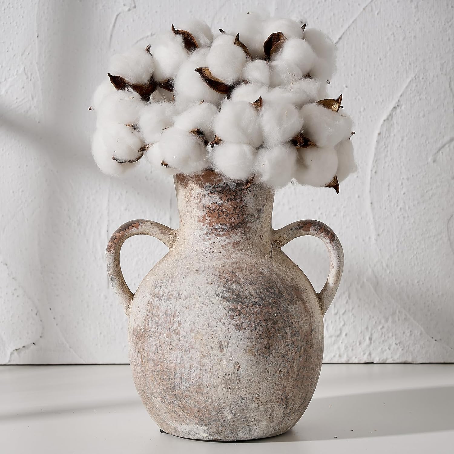 Vintage-style vase with handles holding cotton against a white textured wall.