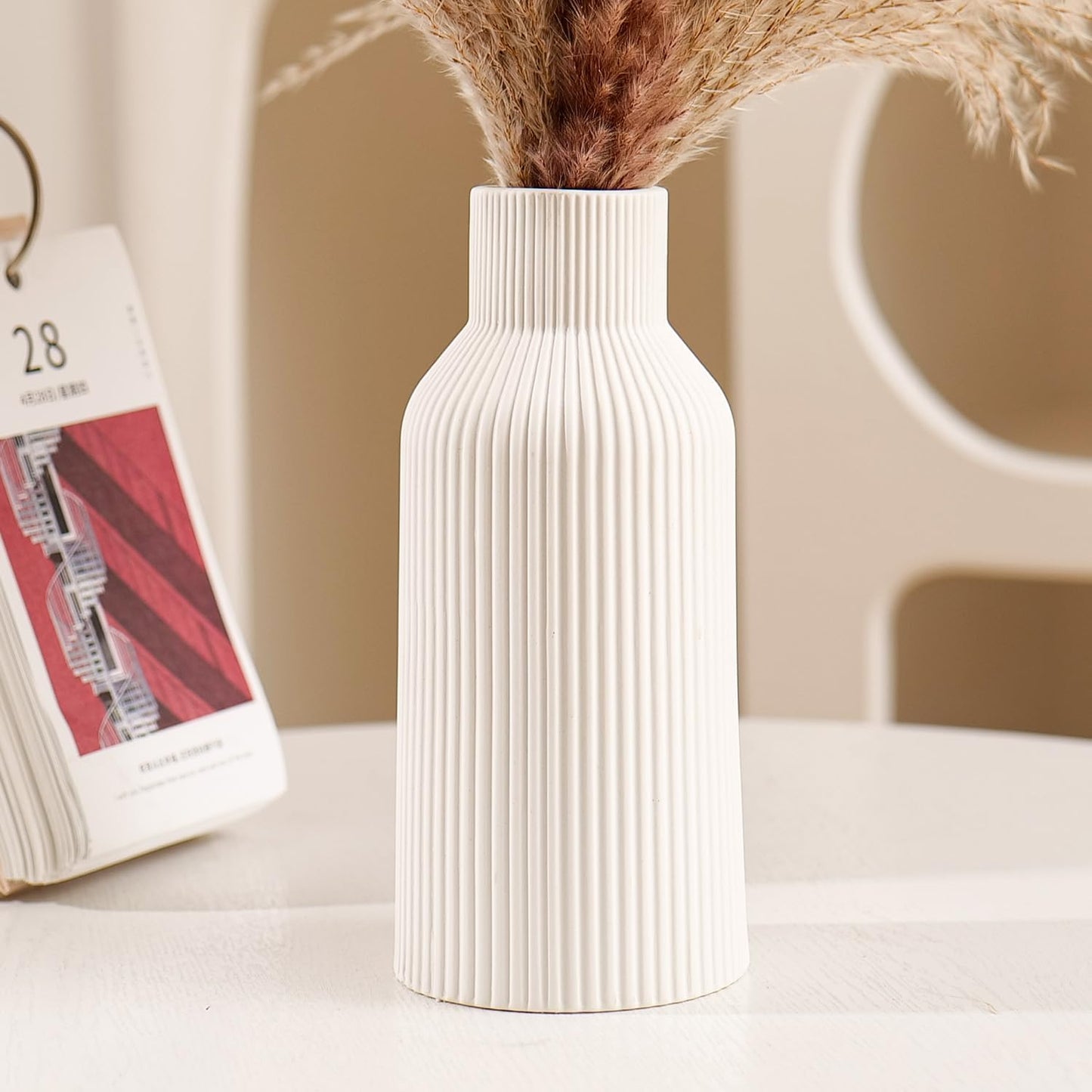 White ribbed vase with dried plants on a light surface, with a blurred calendar in the background.