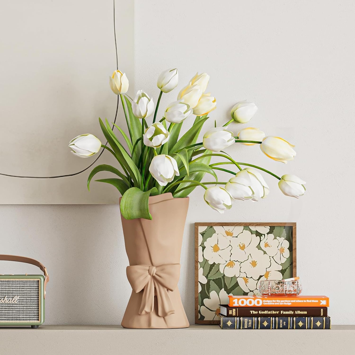 White tulips in a beige vase on a shelf with a framed picture and books in the background.
