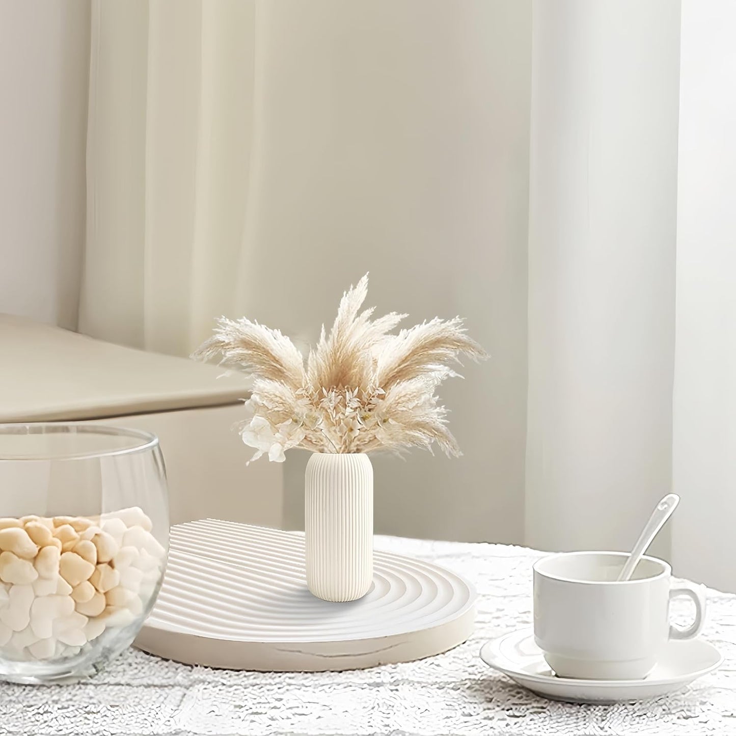 White vase with dried pampas grass on a table with a cup and saucer in a cozy room.