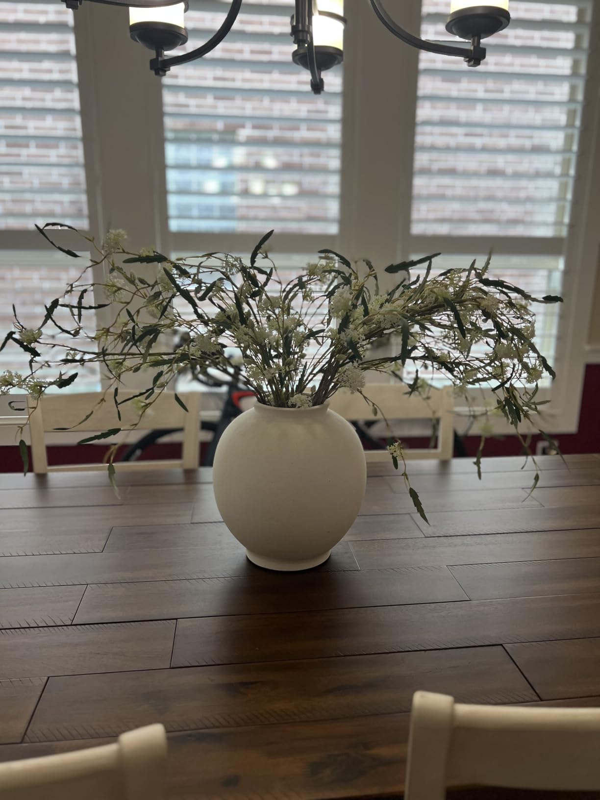 White vase with dried plants on a wooden table in a room with large windows.