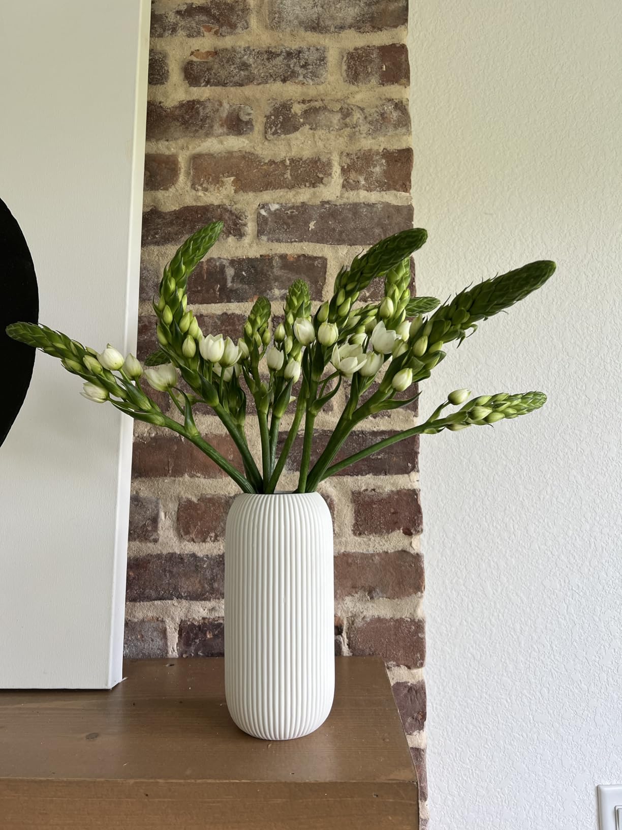 White vase with greenery on a wooden surface against a brick wall.