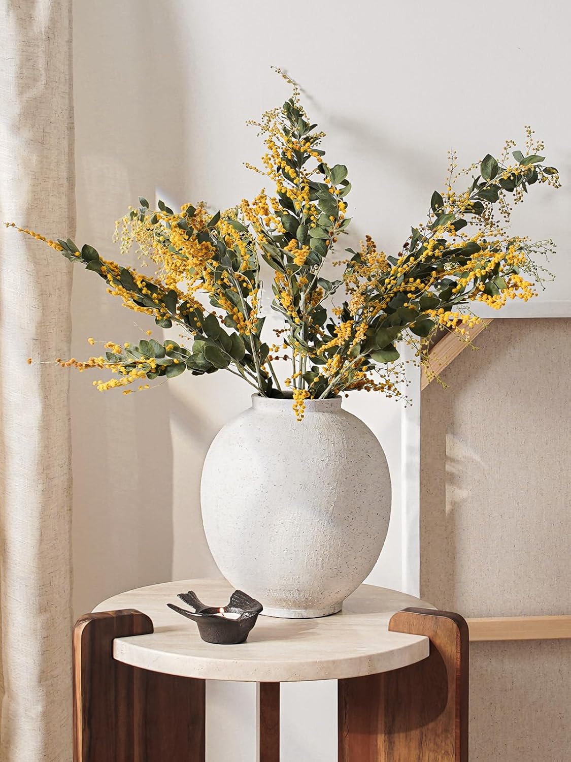 White vase with yellow flowers on a small round table against a neutral wall.